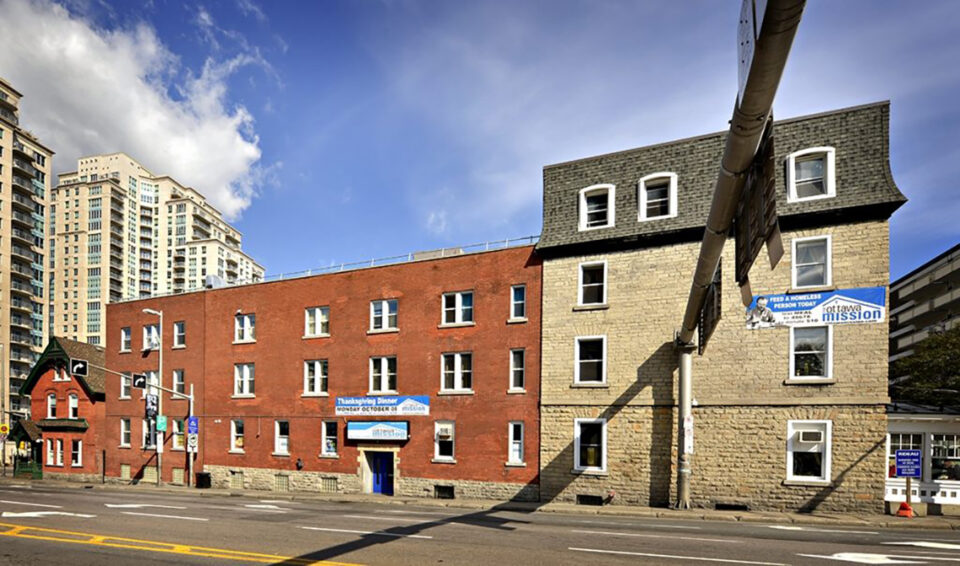 Street view of a red brick building with a sign reading "Open 24 hours a day, 365 days a year." It's sunny, and the street is empty.