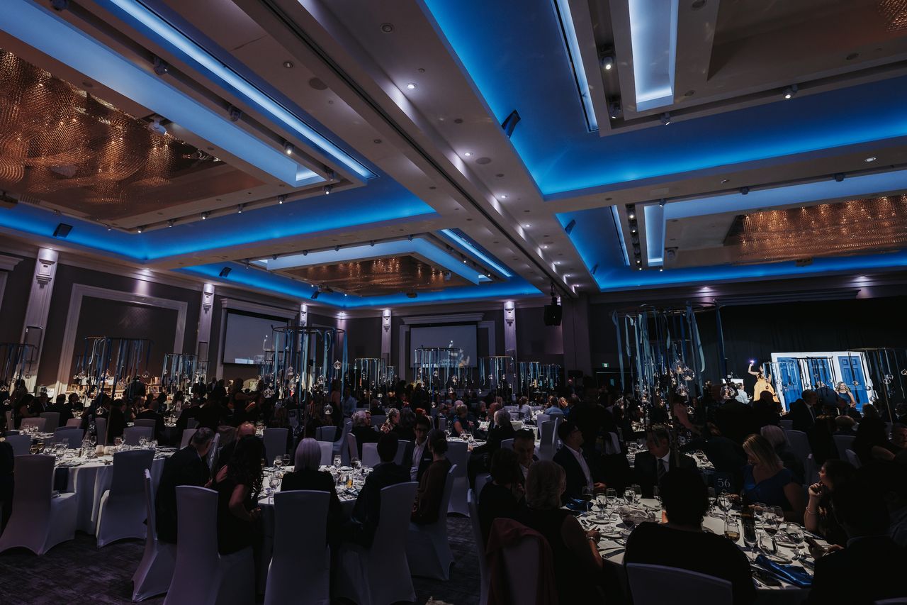 A large banquet hall filled with elegantly dressed guests seated at round tables, under blue-lit ceilings. The ambiance is formal and festive.