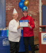 A woman in a white chef's uniform stands smiling, holding a graduation certificate.