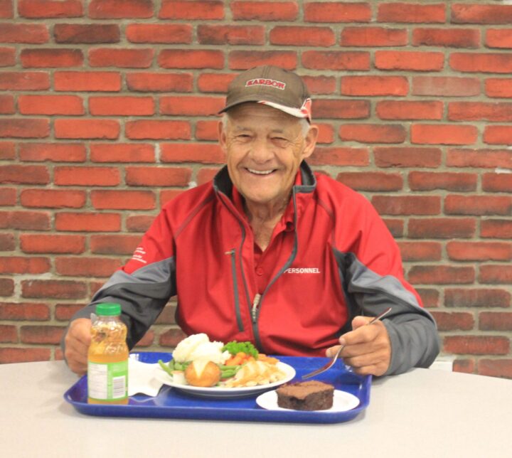 An elderly man is smiling, sitting at The Mission's dining room table with a full meal.