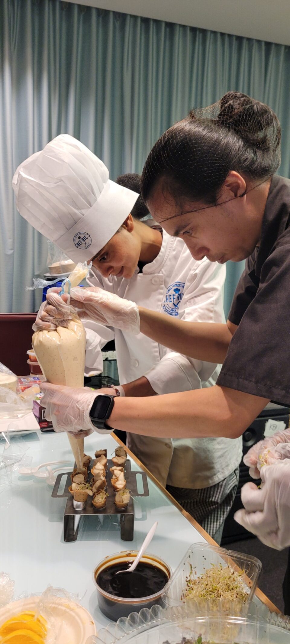 A cook wearing his uniform is showing a student (also wearing her full attire) how to prepare canapes for an event.
