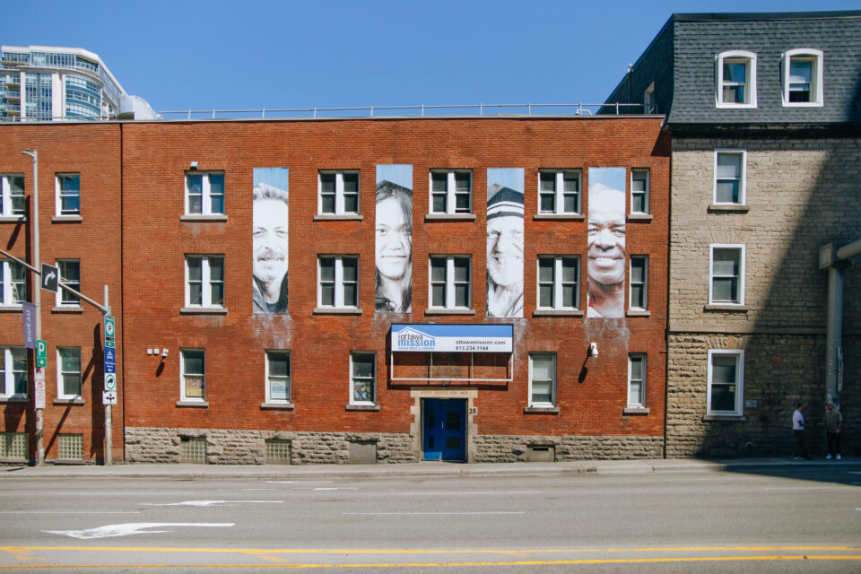 An exterior shot of The Ottawa Mission building: brick building facade showcases large black-and-white portraits between windows of clients, conveying a sense of community and diversity under a clear blue sky.