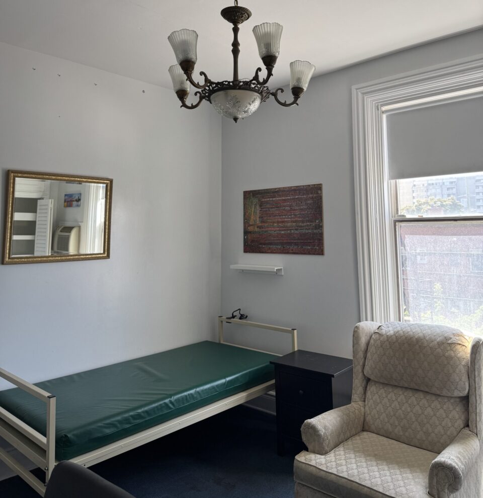 The photo shows the inside of a room at LifeHouse - it’s a minimalist room featuring a vintage chandelier, a single bed with a green mattress, an upholstered armchair, a wall mirror, and a large window letting in natural light.