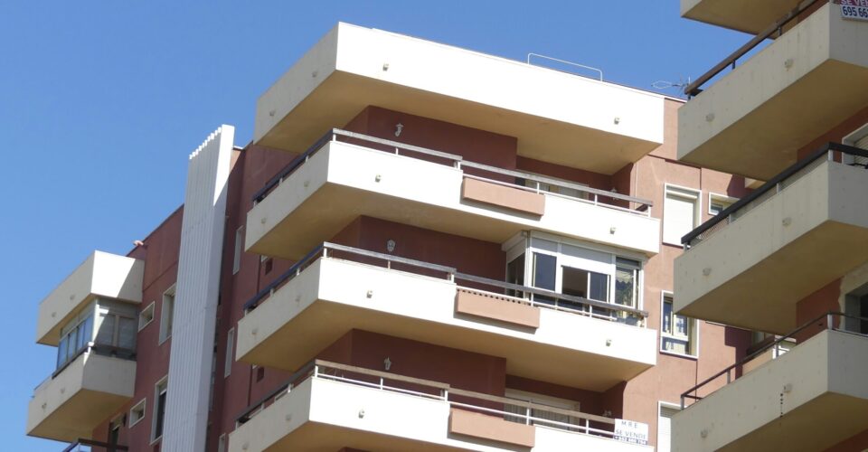 Modern apartment buildings with multiple balconies under a clear blue sky. Beige and brown facades create a clean, geometric look.