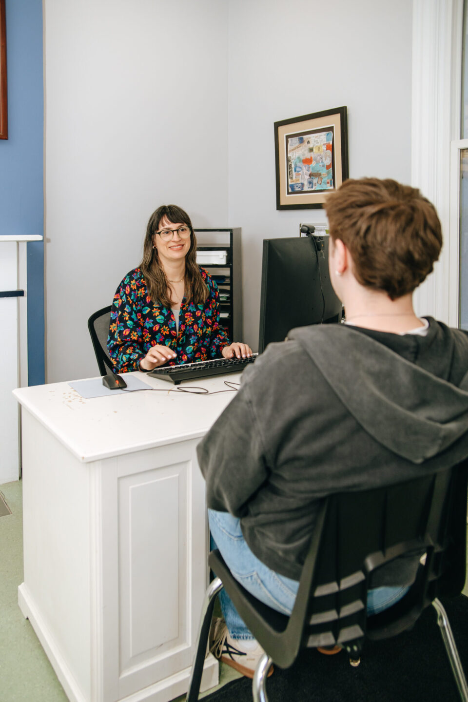 A woman working at The Ottawa Mission wears a colorful blouse and sits at a desk with a computer, smiling at a man in a hoodie. The setting is an office with a calm, professional atmosphere.