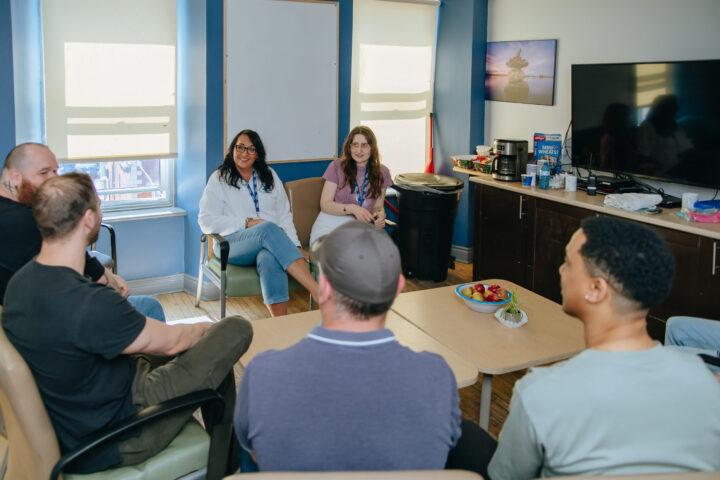 A group of people sit in a circle in a room, receiving counselling, engaged in conversation.