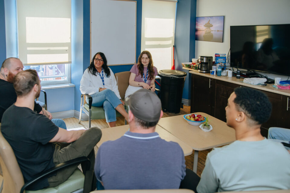 A group of people sit in a circle in a room, receiving counselling, engaged in conversation.