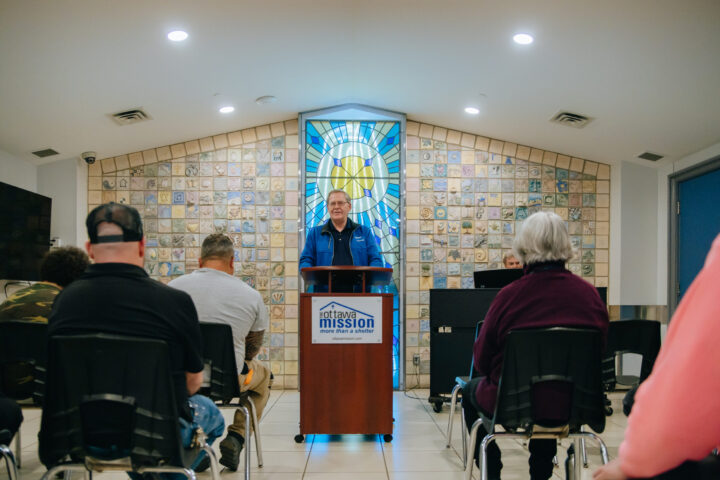 A speaker stands at a podium labeled "Ottawa Mission" inside a room with a colorful stained-glass window. Seated audience members listen attentively.