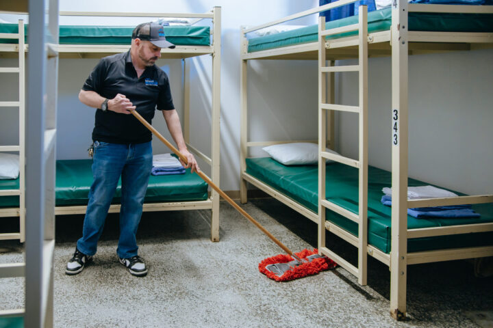 Man mopping a clean, orderly room with bunk beds and green mattresses.