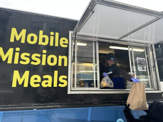 A chef in a "Mobile Mission Meals" truck hands a food bag to a person outside.