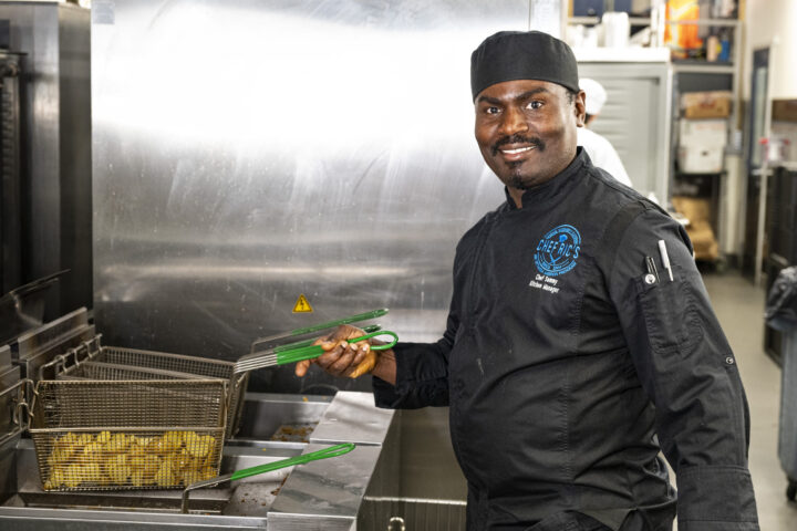Chef in black uniform and cap smiles while holding green tongs, frying potatoes in a commercial kitchen.
