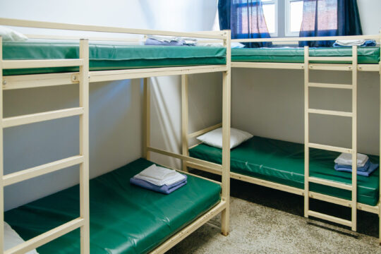 A Mission dorm room with two sets of bunk beds featuring green mattresses, a window with blue curtains, and neatly folded towels and pillows on each bed.