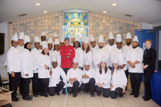 Group photo of Food Services Training Program (FSTP) in white uniforms and chef hats, surrounding a chef in a red jacket. They pose happily in front of a stained glass window in the Mission chapel.
