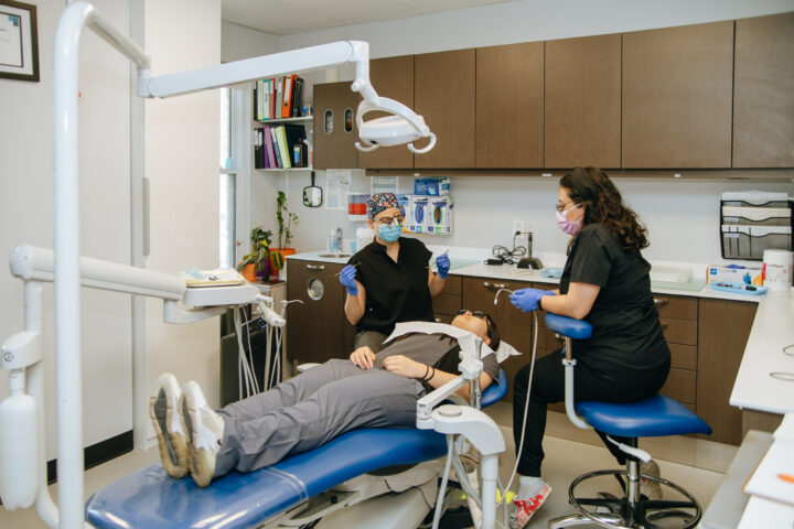 Dentists in masks and gloves attend to a patient reclining in a dental chair. The office is organized and equipped with dental tools.