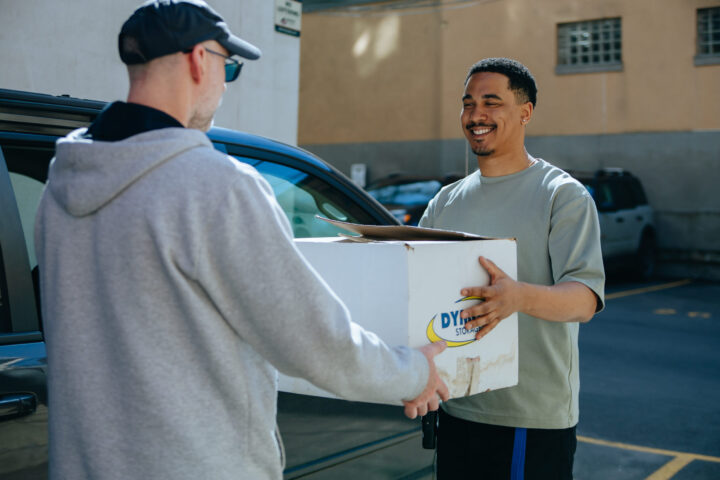 Two men exchange a large white box outdoors near a parked car. The man on the right smiles warmly, creating a friendly and cooperative atmosphere.