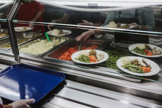 A cafeteria scene with gloved hands serving meals. Plates contain mashed potatoes, green beans, sliced carrots, turkey, and stuffing. A blue tray is held below.