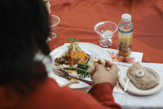 An Ottawa Mission guests enjoys a meal with turkey, carrots, and green beans on a white plate in the dining room. Nearby are a dessert, bottle drink, and empty cups on a fall-themed tablecloth.