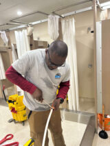 Abraham wears a gray shirt and works on a white pipe in a room with shower stalls and yellow cleaning equipment.