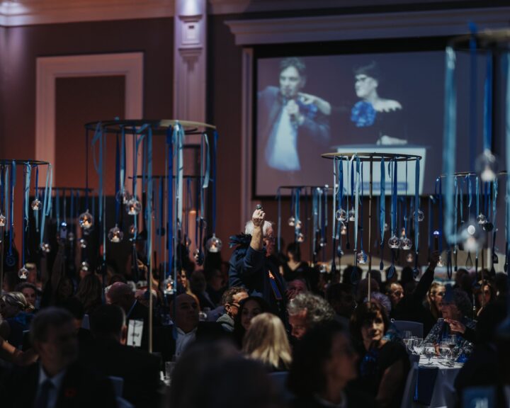 Blue Door Gala - guests gather at tables crowded by attendees, decorated with hanging blue ribbons and glass ornaments. A projector displays a speaker on stage.