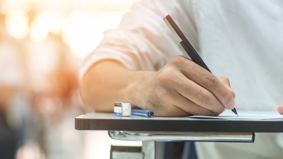 A person writes with a pen on paper at a school desk. An eraser and another pen rest nearby. The setting is bright, conveying focus and study.