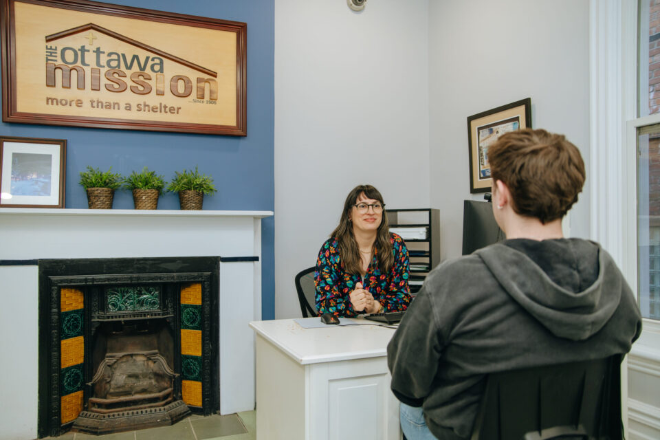 A woman with glasses and a floral top sits at a desk, smiling at a man in a hoodie. A sign reads "Ottawa Mission." The room feels welcoming and supportive.