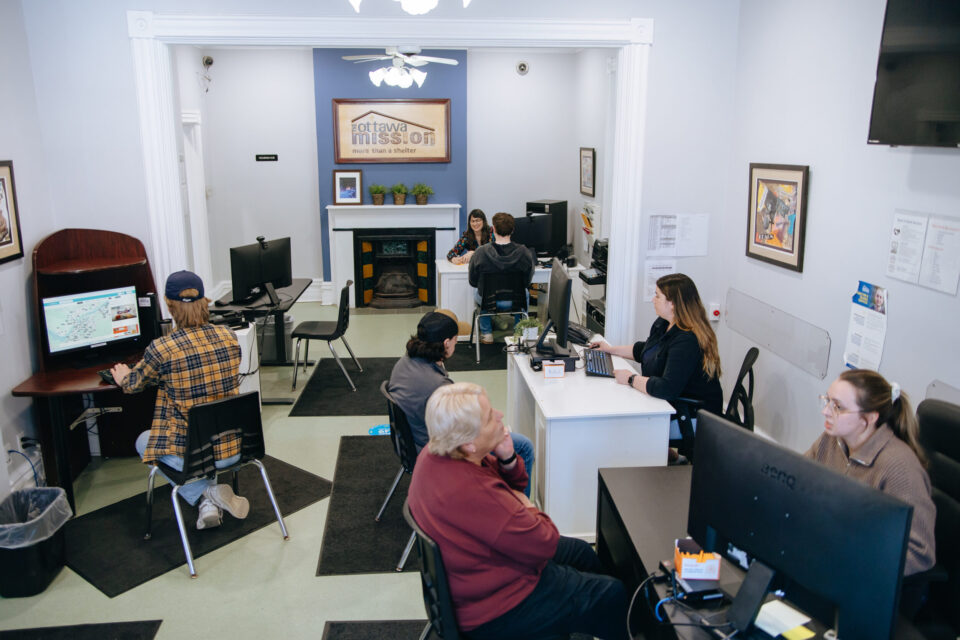 Various people sit at desks inside a well-lit, clean room. A sign at the back of the room reads: "The Ottawa Mission."