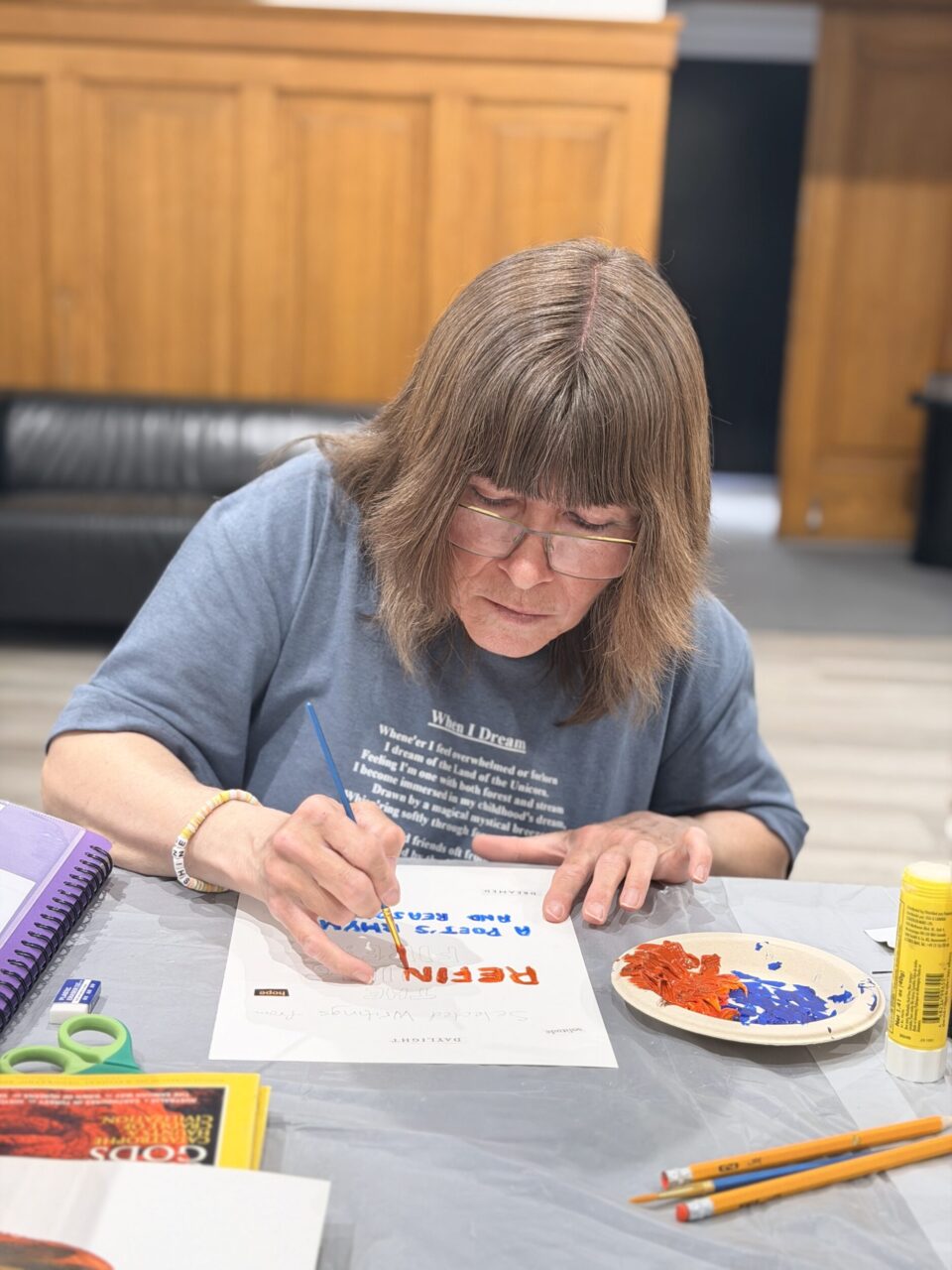 A woman with brown hair and glasses paints words onto a piece of paper.