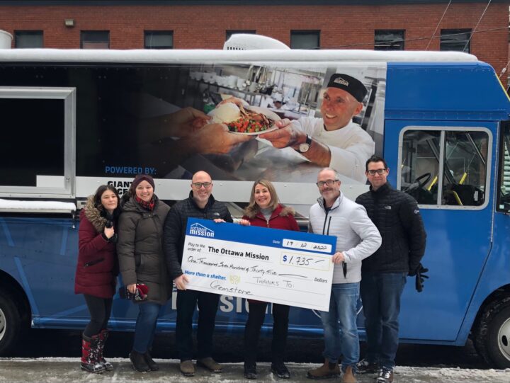 Group of six smiling people holding a large donation check in winter attire, standing before a blue Ottawa Mission food truck with a Chef Ric’s image on it.
