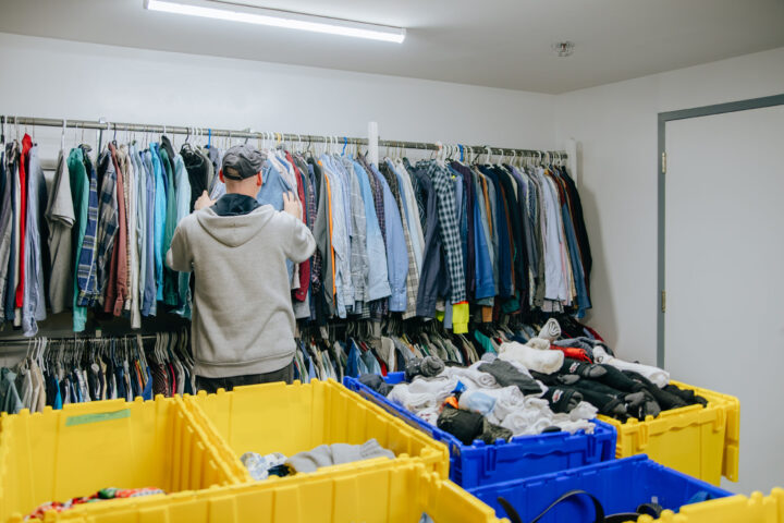 A Ottawa Mission client in a hoodie and cap looks through clothes on a rack. Yellow bins filled with clothes are in the foreground.