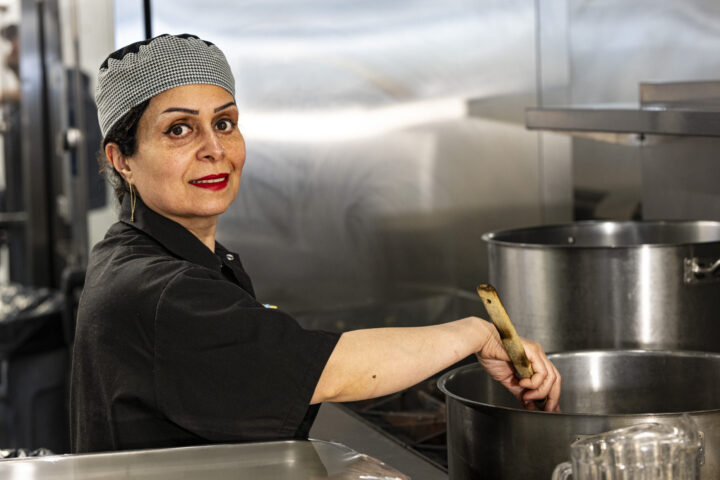 A chef in a black uniform and checkered cap stirs a large pot with a wooden spoon in The Mission kitchen, looking at the camera with a slight smile.