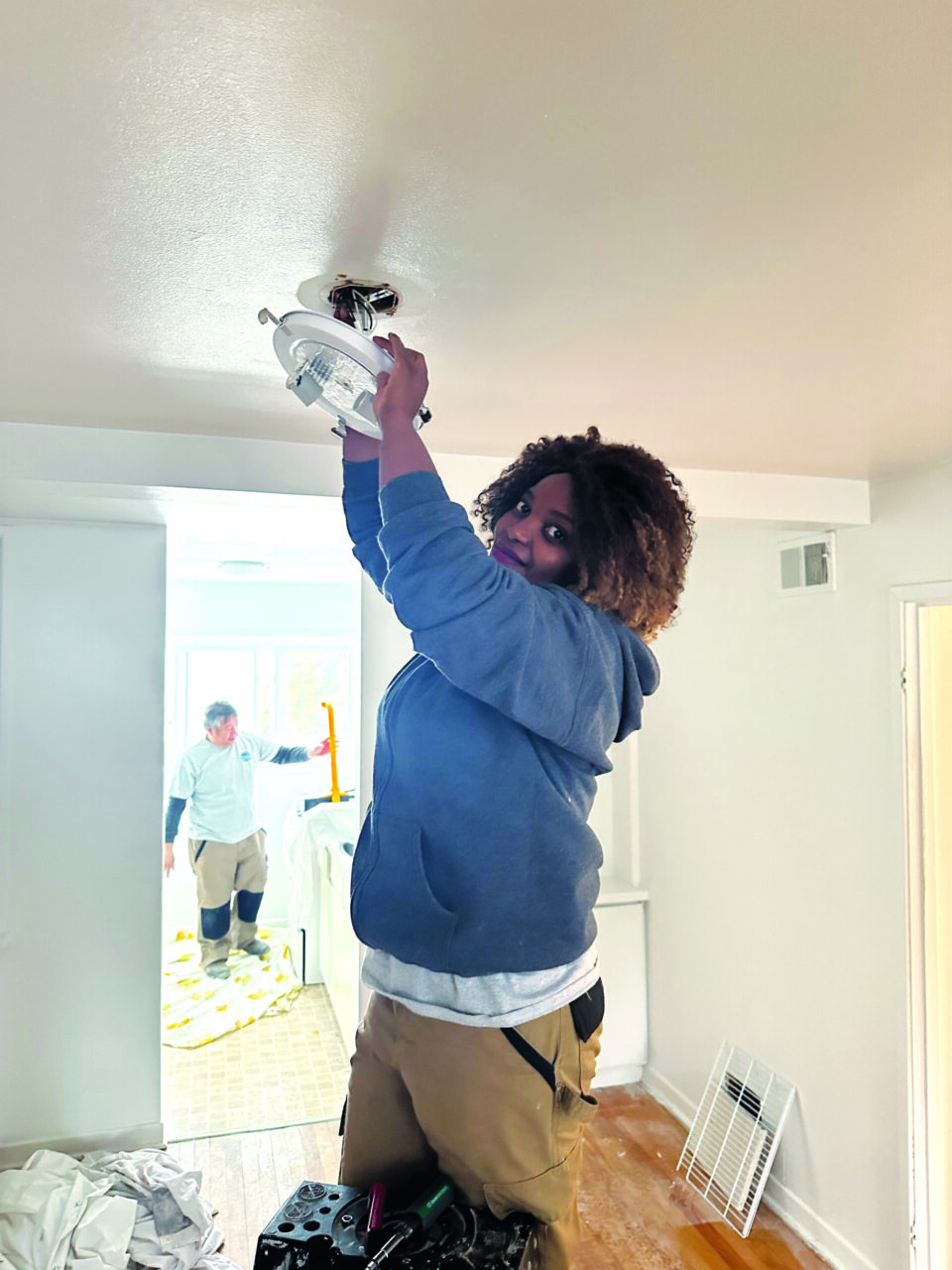 Gloria wears a hoodie smiling while installing a ceiling light in a bright room.