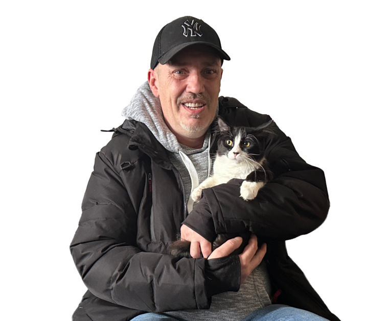 John sits in a black jacket and cap sitting on a black sofa, smiling and holding a black-and-white cat.