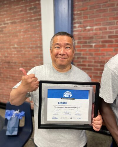 Leo man smiling and holding a MSTP graduation certificate with a thumb up. He stands in front of a red brick wall, conveying pride and achievement.