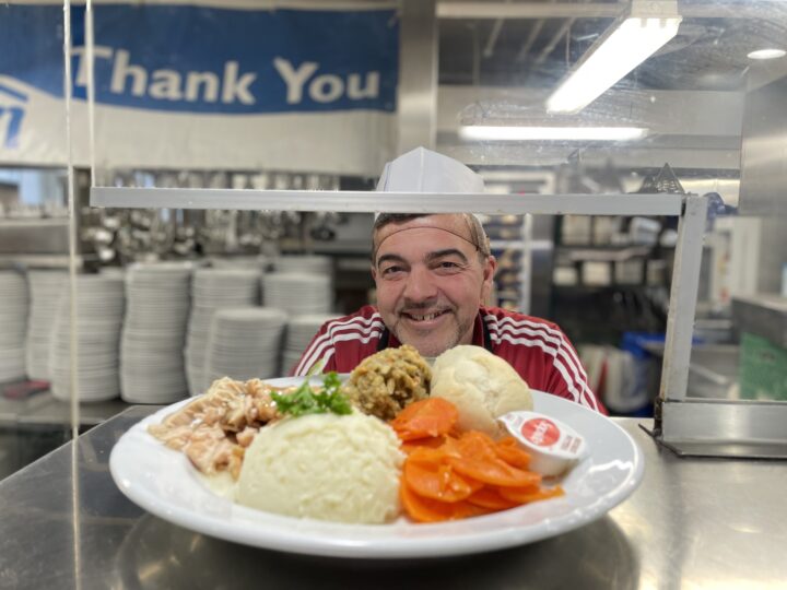Smiling man behind The Mission kitchen counter presents a holiday thanksgiving dinner. of mashed potatoes, carrots, A "Thank You" sign is visible in the background.