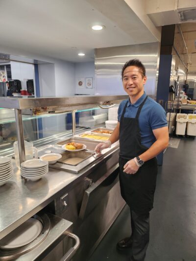 Smiling man in apron serves food at a cafeteria line. Plates and trays of food are visible, indicating a welcoming and busy atmosphere.