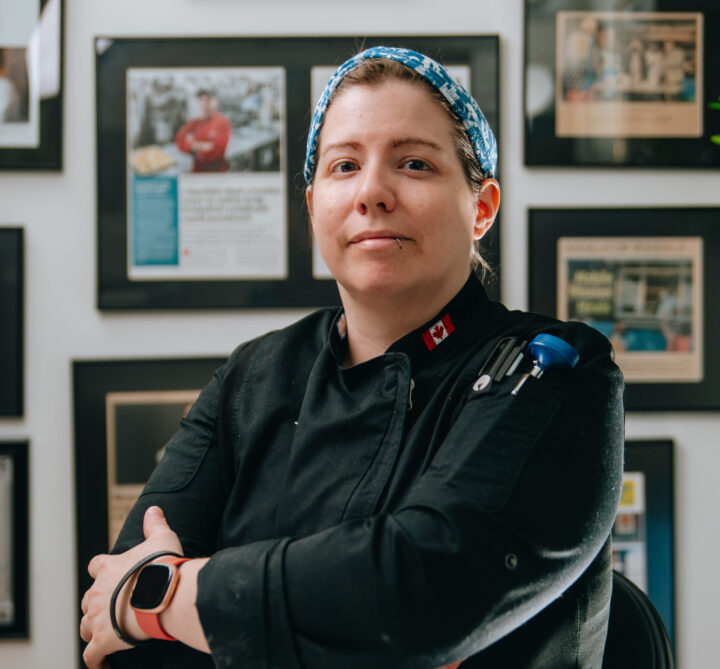 Chef Jennifer stands in a black uniform with folded arms, wearing a blue headscarf. Background features framed accolades, conveying professionalism and confidence.