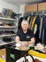 A person with short hair smiles while sorting clothes in a storage room. Surrounding shelves are filled with shoes, and colorful clothes hang on a rack.