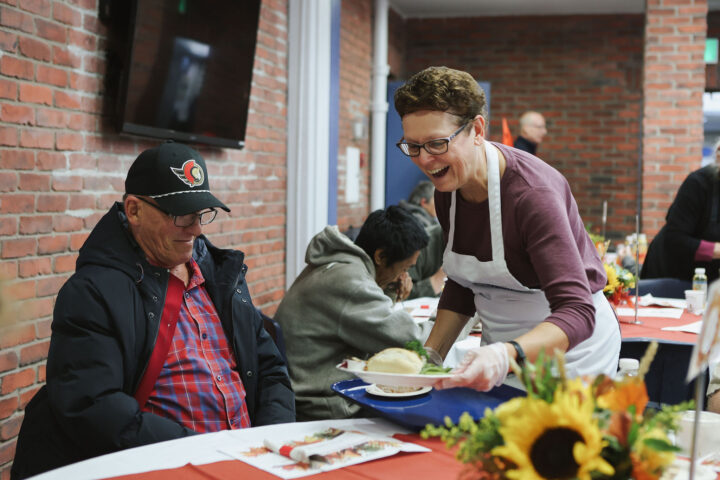 A smiling woman in an apron serves a meal to a seated man wearing a cap at a community center with brick walls, decorated with sunflowers.