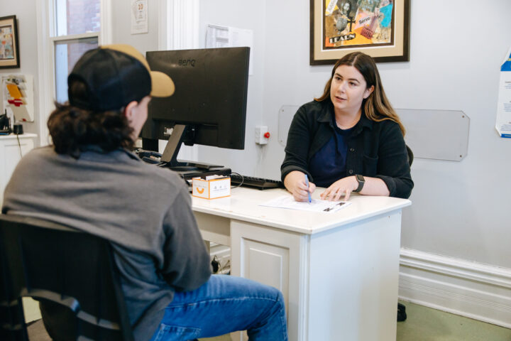 A woman at a desk attentively listens to a man with a cap, taking notes. The office setting is casual, and the atmosphere is focused yet relaxed.