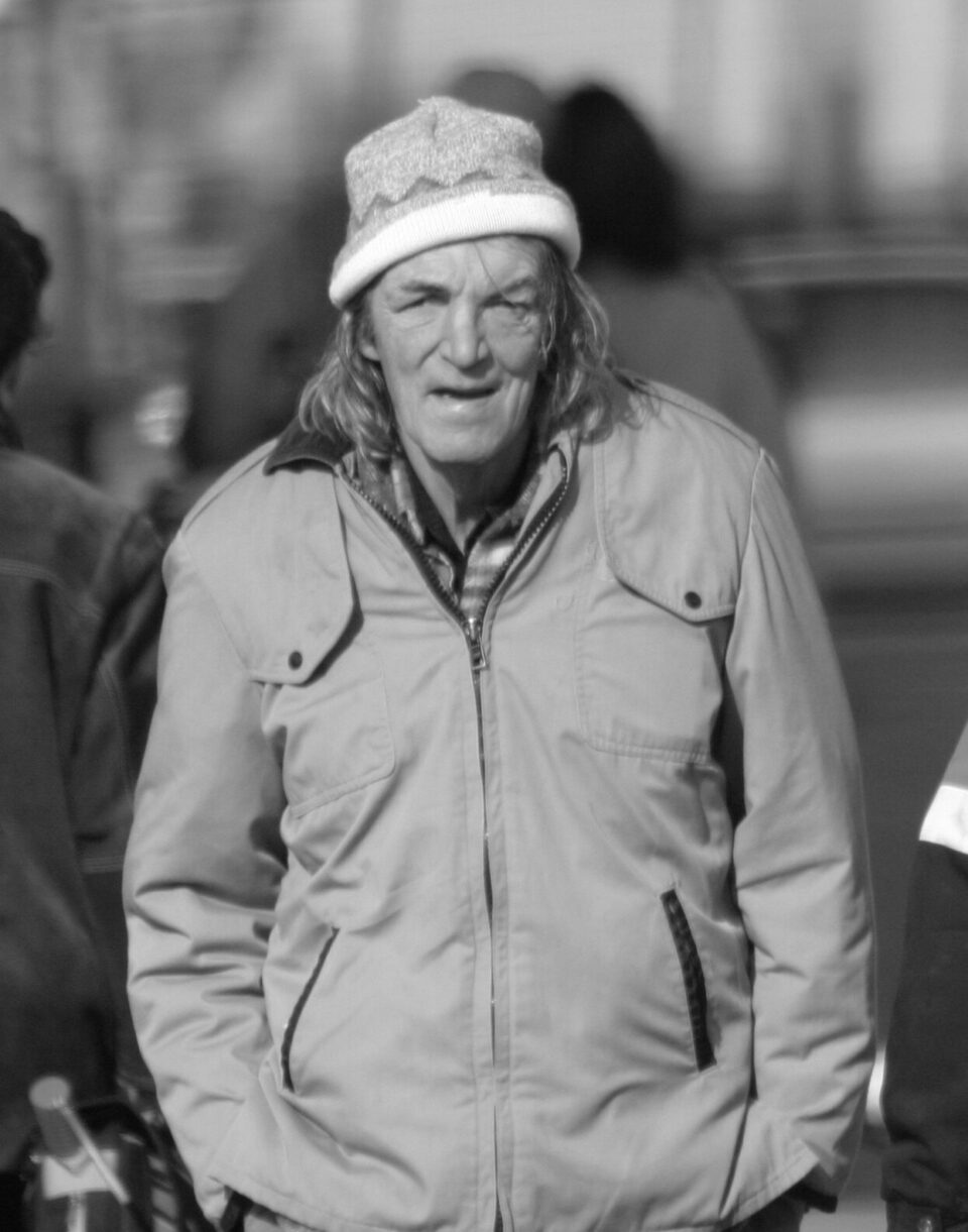 Black & white photo. Elderly person with long hair and a hat, wearing a coat, walks outdoors.