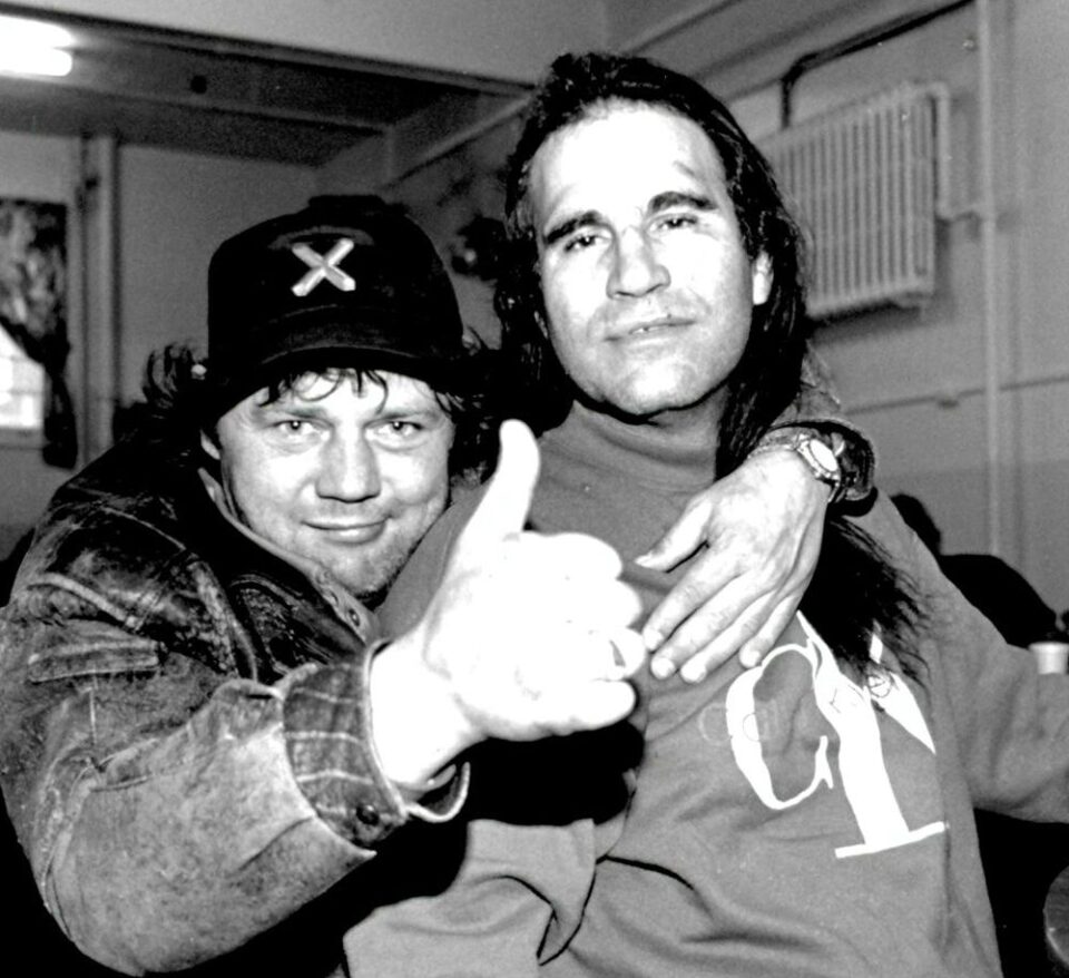 wo men pose closely for the camera; one smiles broadly, giving a thumbs-up, while the other has a relaxed expression. The setting appears casual.