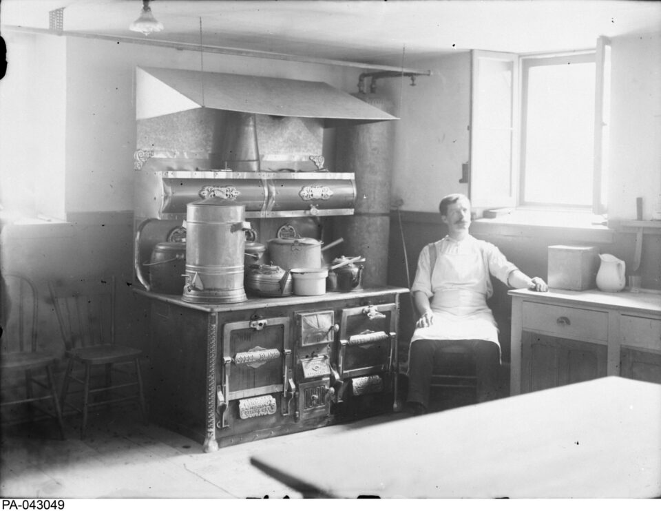 Black and white photo of a man in an apron sitting beside a vintage stove in a traditional kitchen, conveying a sense of nostalgia and calm.