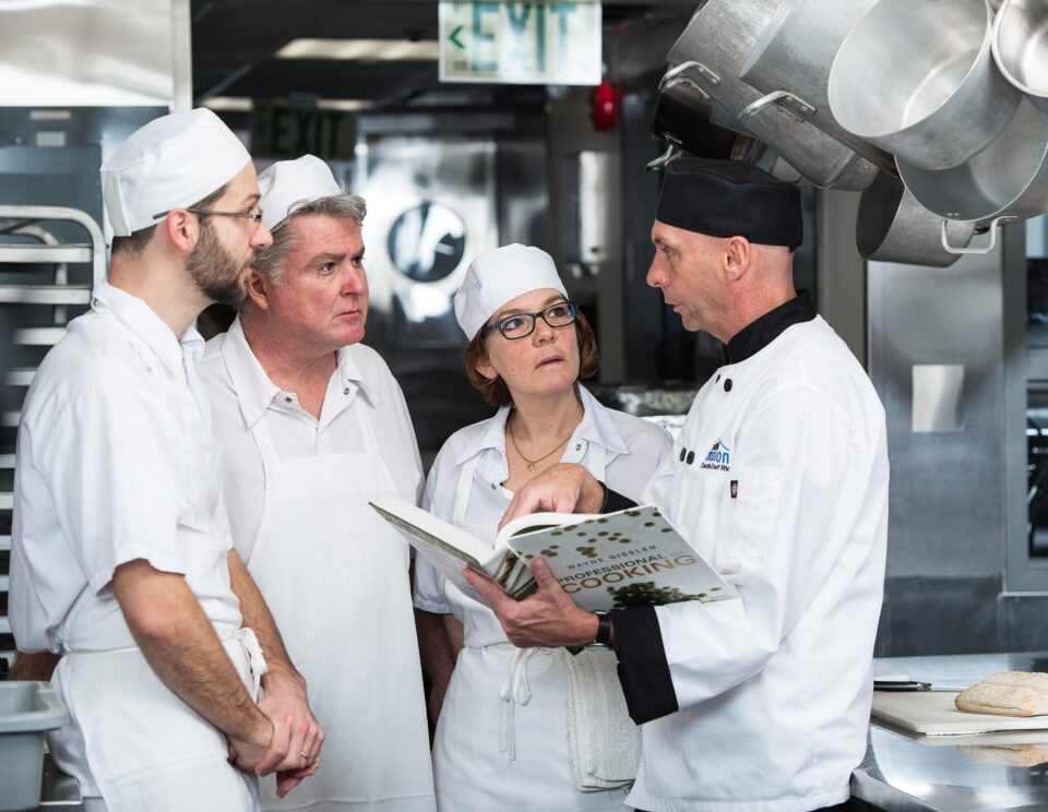 Chef Rics is wearing a black hat discusses a recipe with three sous chefs in a professional kitchen. They are focused on an open cookbook.