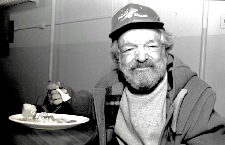 Black and white photo of a bearded man in a hoodie and cap smiles while holding a fork beside a plate of food, eating at The Ottawa Mission .