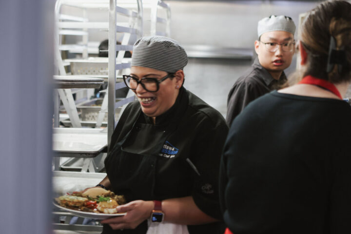 A smiling chef in glasses carries a plated dish in a busy kitchen, wearing a striped cap and black uniform. The atmosphere is lively and bustling.