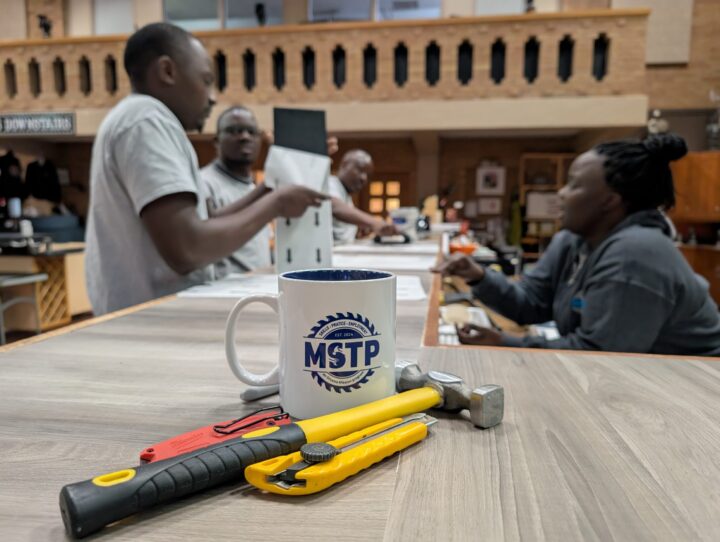 A mug with the MSTP logo sits on a wooden table alongside tools. In the blurred background, people engage in discussion, creating a collaborative atmosphere