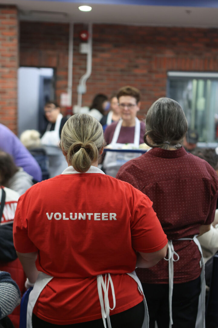 Volunteer in a red shirt with "VOLUNTEER" printed on the back, stands in a busy soup kitchen. Others in aprons serve meals, conveying a sense of community and teamwork.