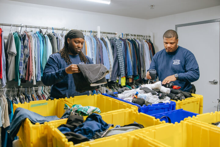 Two frontline employee sorting clothes in The Ottawa Mission’s clothing room.. Yellow bins filled with clothes and shelves of hanging garments surround them.