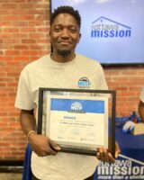 Ronalad smiles while holding a framed Maintenance Services Training Program certificate. He's wearing a gray MSTP t-shirt. A "The Ottawa Mission" logo is visible on a brick wall behind him, creating a proud and celebratory atmosphere.