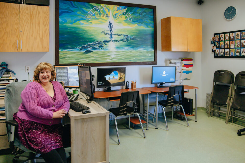 A woman in a pink sweater smiles while sitting at a desk with a computer. Behind her, a bright mural and two computer stations are visible in a cozy classroom.